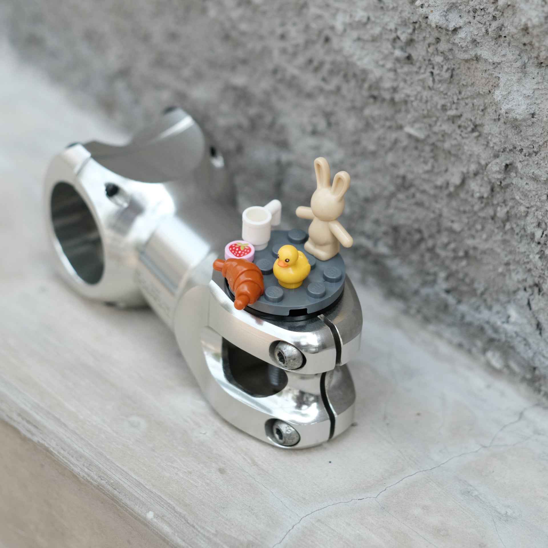 A silver metal bicycle stem clamp with a small decorative miniature table setup on top, placed on a light stone surface against a textured gray concrete wall.